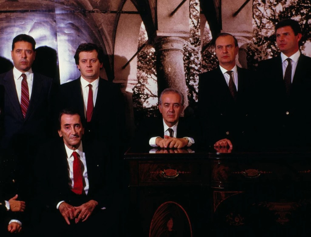 Photograph entitled "The Financiers" by Michael Clegg & Martin Guttmann, picturing six men in suits and and red ties, and stoic faces, seeming to pose for a group portrait.